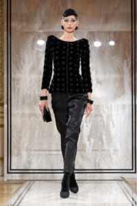 Female model on a runway wearing a black velvet jacket with circular embellishments, matching black satin-like pants, and a black cap with bold earrings and bracelets, holding a small clutch