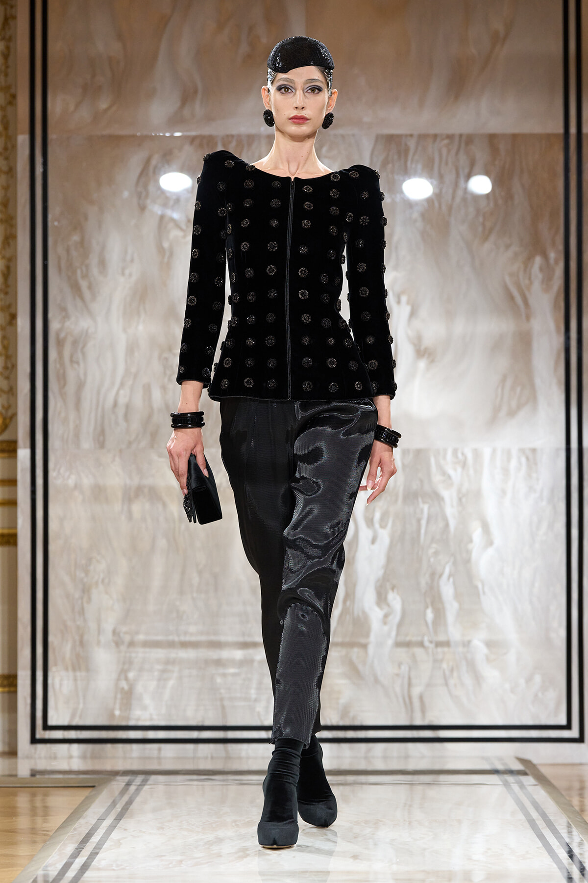 Female model on a runway wearing a black velvet jacket with circular embellishments, matching black satin-like pants, and a black cap with bold earrings and bracelets, holding a small clutch