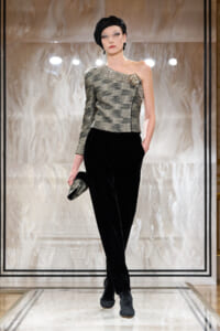 Model on a runway wearing a metallic one-shoulder top with black velvet trousers, black turban, earrings, and a small metallic clutch.