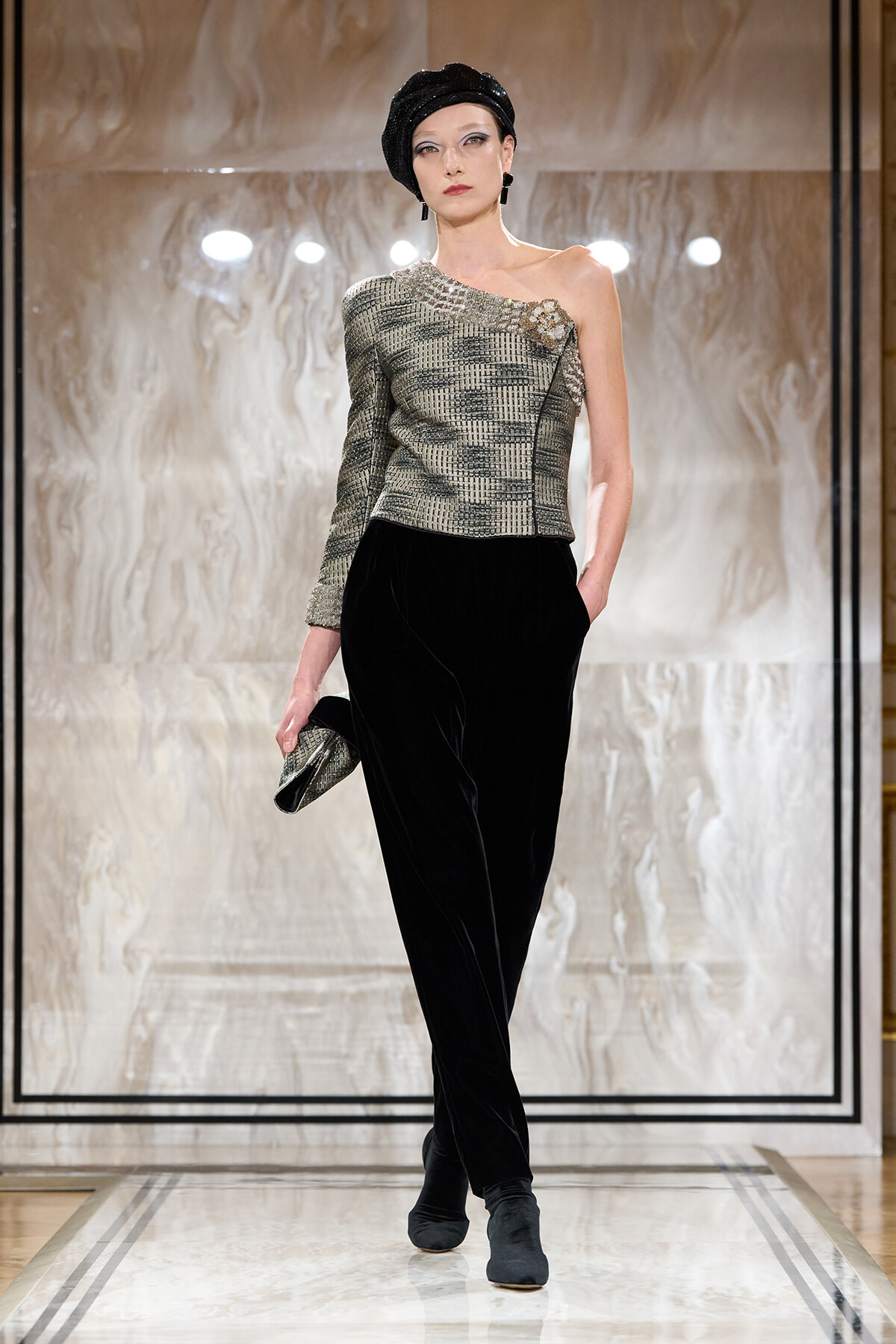 Model on a runway wearing a metallic one-shoulder top with black velvet trousers, black turban, earrings, and a small metallic clutch.