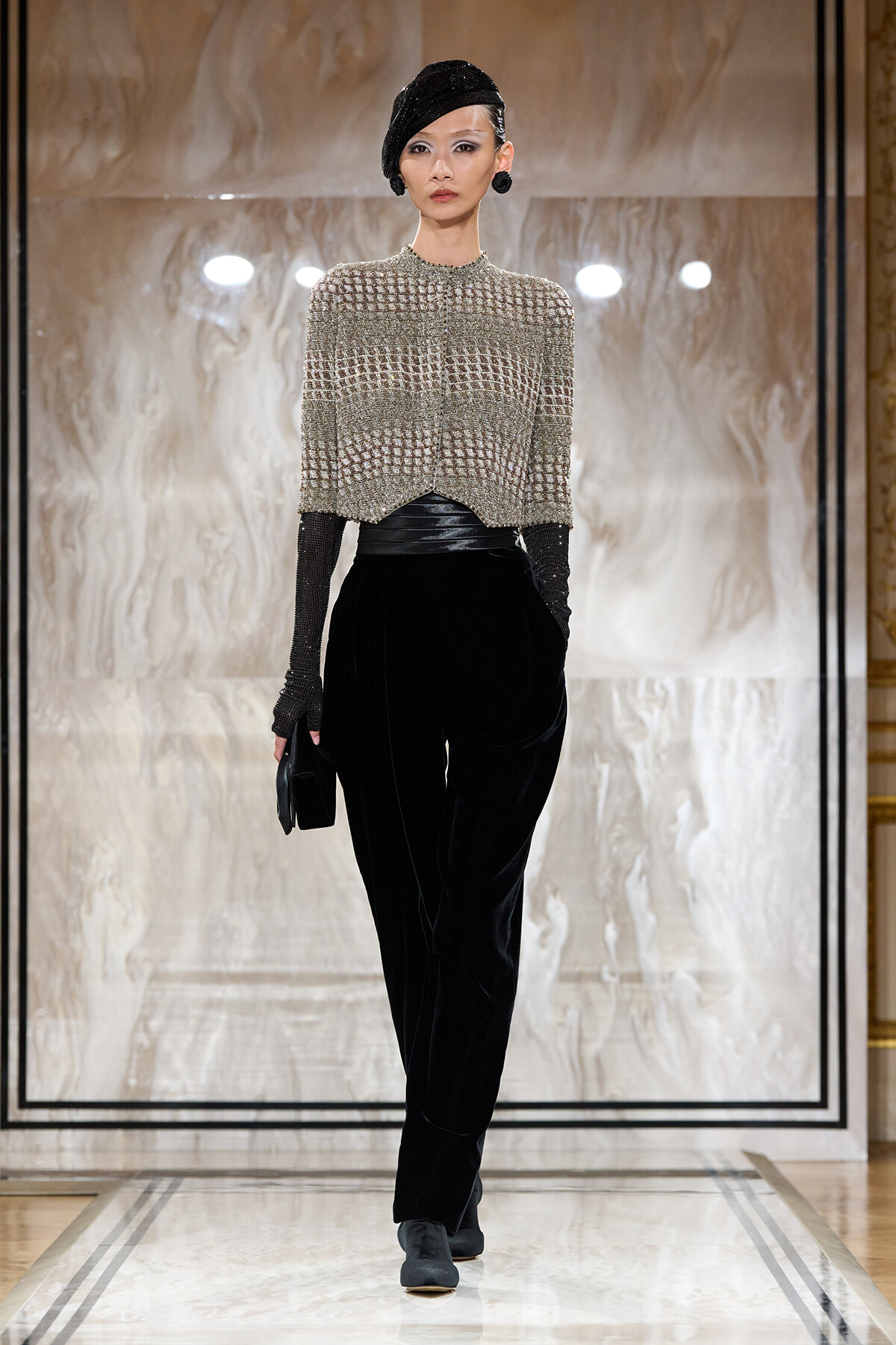 Model walking a runway in a metallic mesh top, black velvet pants, and a black headwrap with large earrings and gloves, holding a small black clutch.