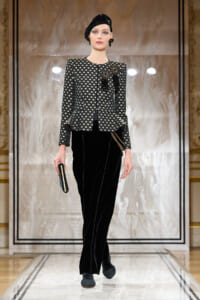 Model on a fashion runway wearing a black patterned cropped jacket with a peplum, black velvet wide-leg pants, a black headwrap, and a small clutch against a marble backdrop.
