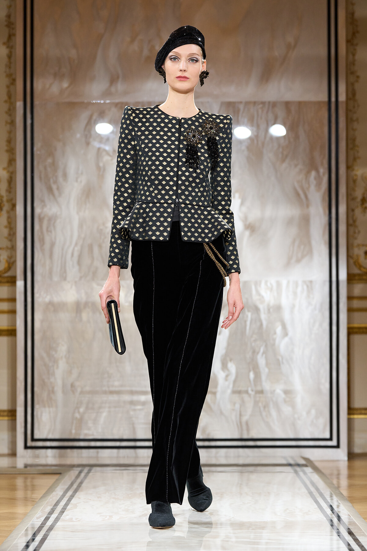 Model on a fashion runway wearing a black patterned cropped jacket with a peplum, black velvet wide-leg pants, a black headwrap, and a small clutch against a marble backdrop.