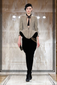 Model on a runway wearing a sparkly silver blazer, black velvet pants, and a black hat, holding a small clutch in her hand.