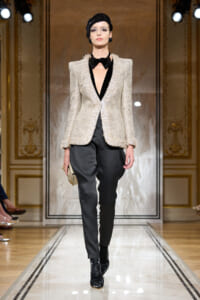 Model walks a runway in a beige textured blazer, black deep-V top, black tapered pants, black ankle boots, and a black headwrap, holding a beige clutch.