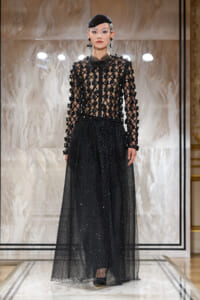 Model on runway wearing an all-black beaded lattice top with a sheer polka-dotted skirt, headpiece and matching accessories.