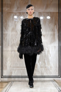 Model on a runway wearing a black sequin top with feather trim, black pants, and a small black headpiece.