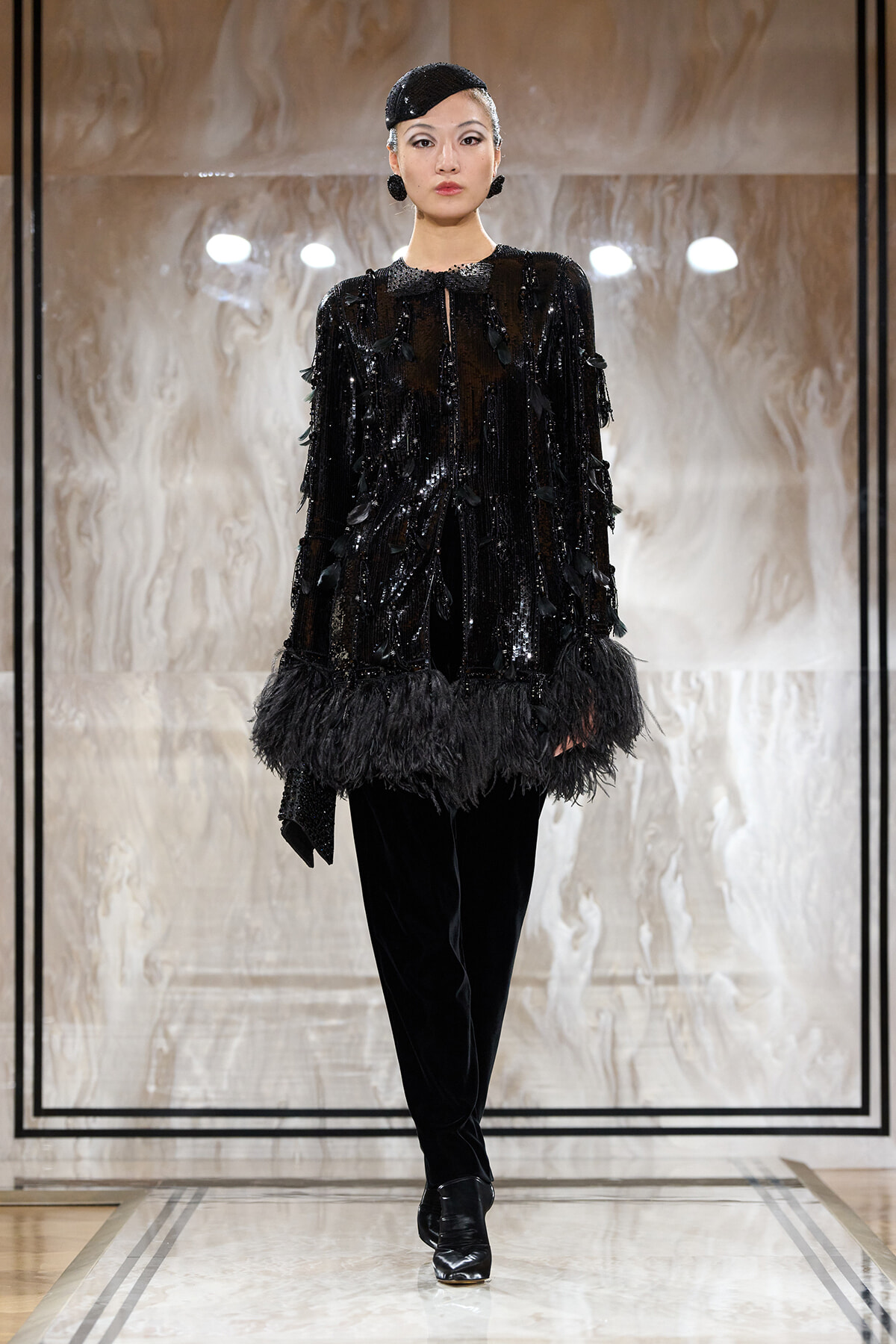 Model on a runway wearing a black sequin top with feather trim, black pants, and a small black headpiece.