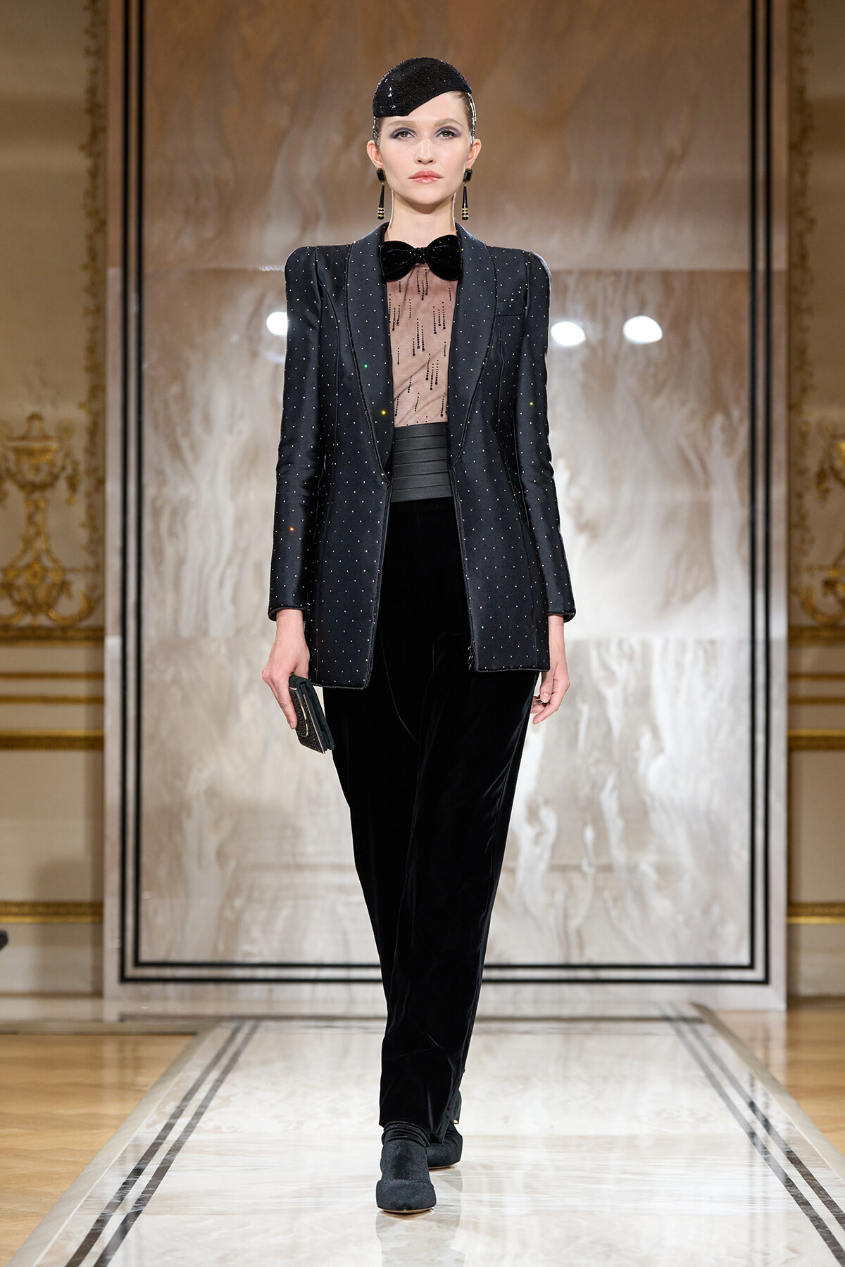Model on a marble runway wearing a sparkly black dotted blazer over a sheer blouse with a bow, paired with black velvet pants and a small clutch in hand.
