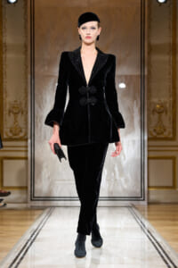 Model walking the runway in a black velvet suit with a deep V neckline, frogged closures, and ruffled cuffs against an ornate backdrop.
