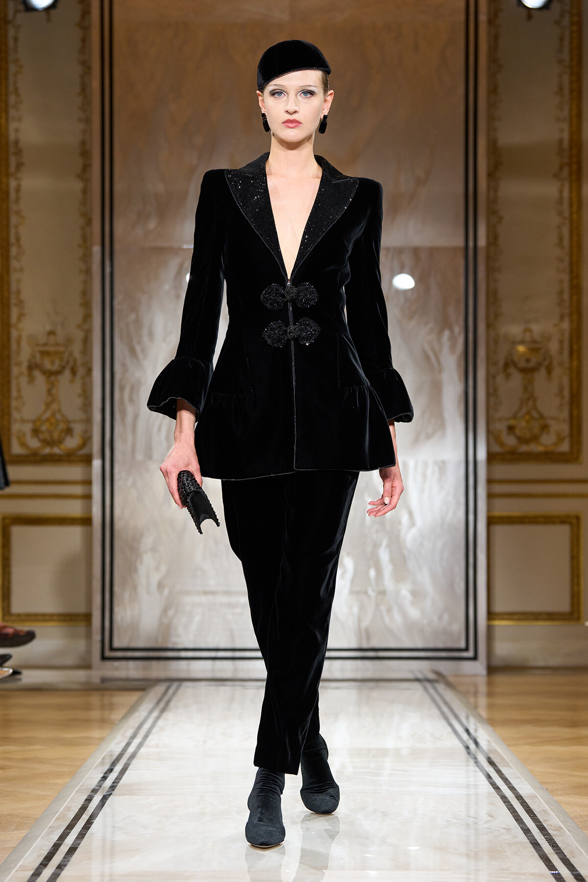 Model walking the runway in a black velvet suit with a deep V neckline, frogged closures, and ruffled cuffs against an ornate backdrop.