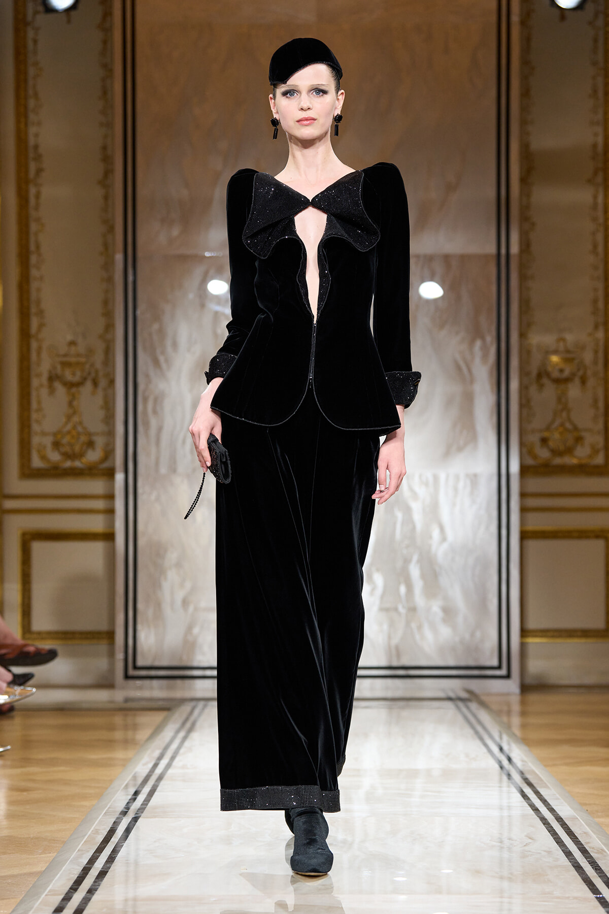 Model walking a runway in a black velvet suit with an oversized bow neckline and matching accessories against a gilded backdrop.