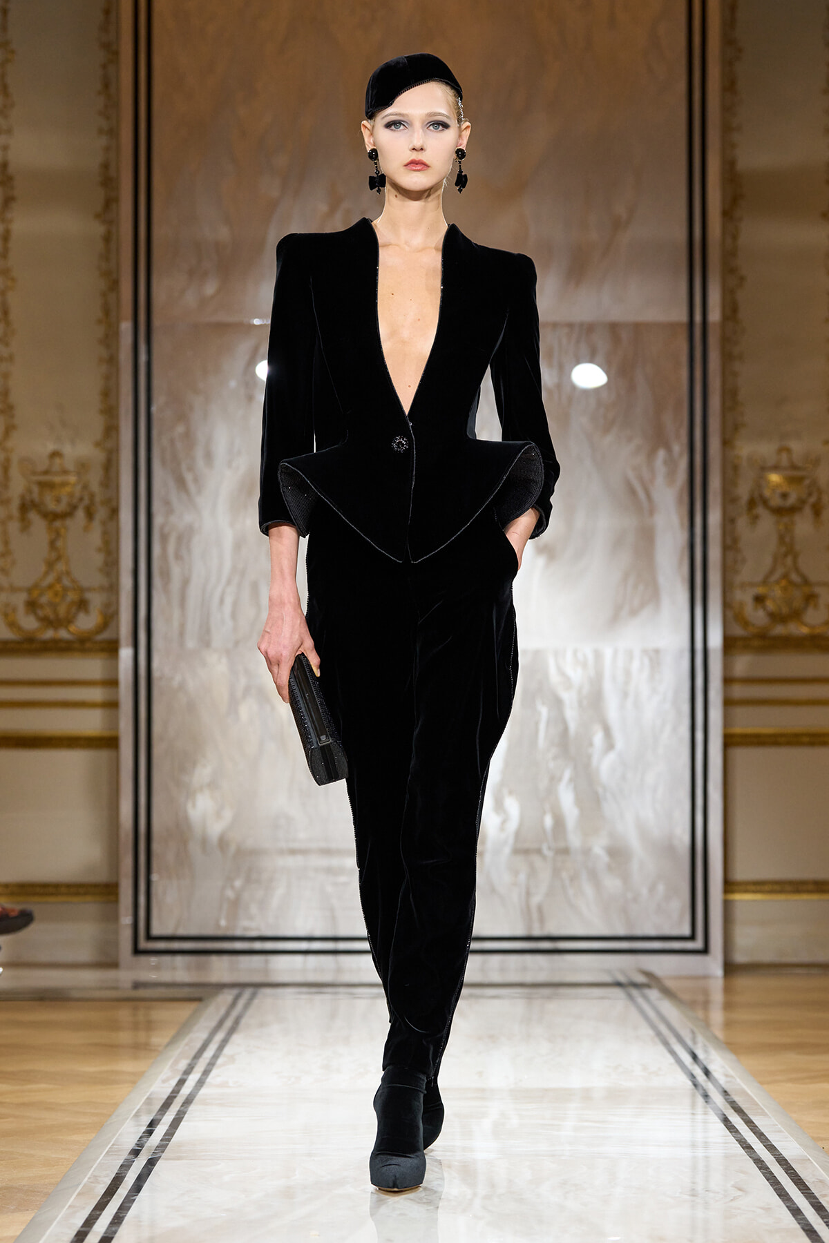 Model in a black velvet blazer and matching pants walks the runway, hands in pockets.