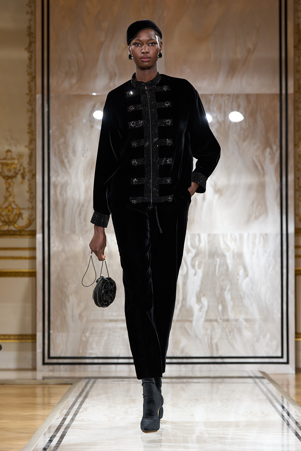 Runway model in a black velvet military-style jacket with silver buttons and matching pants, carrying a small round black handbag.