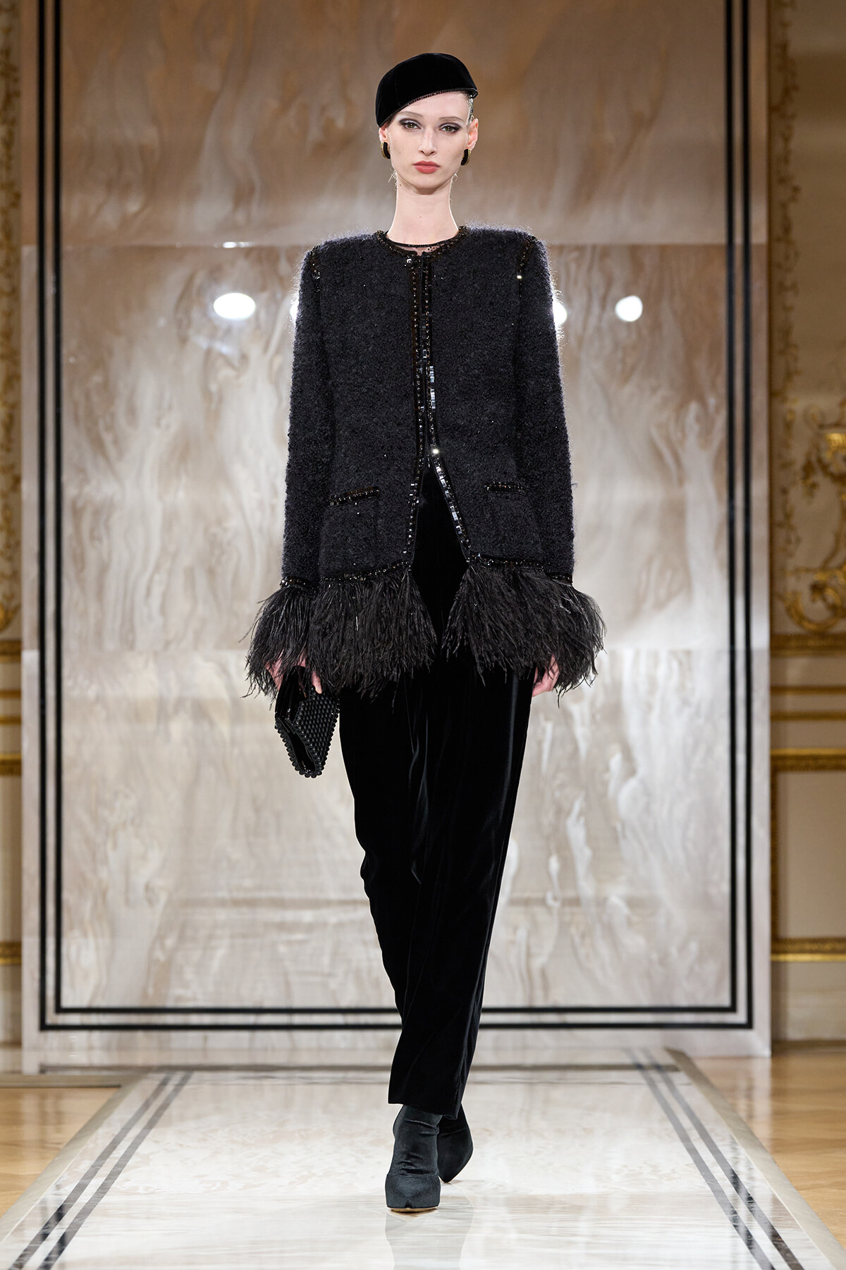 Model on a runway wearing a black textured coat with feathered hem, black velvet pants, and a beret, holding a clutch.