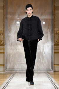 Model walking a fashion show in a textured black cardigan and velvet wide-leg pants, accessorized with a black hat and chunky earrings.