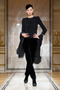 Model struts a runway in a black textured coat with fringe cuffs, matching velvet pants, and a small black cap.