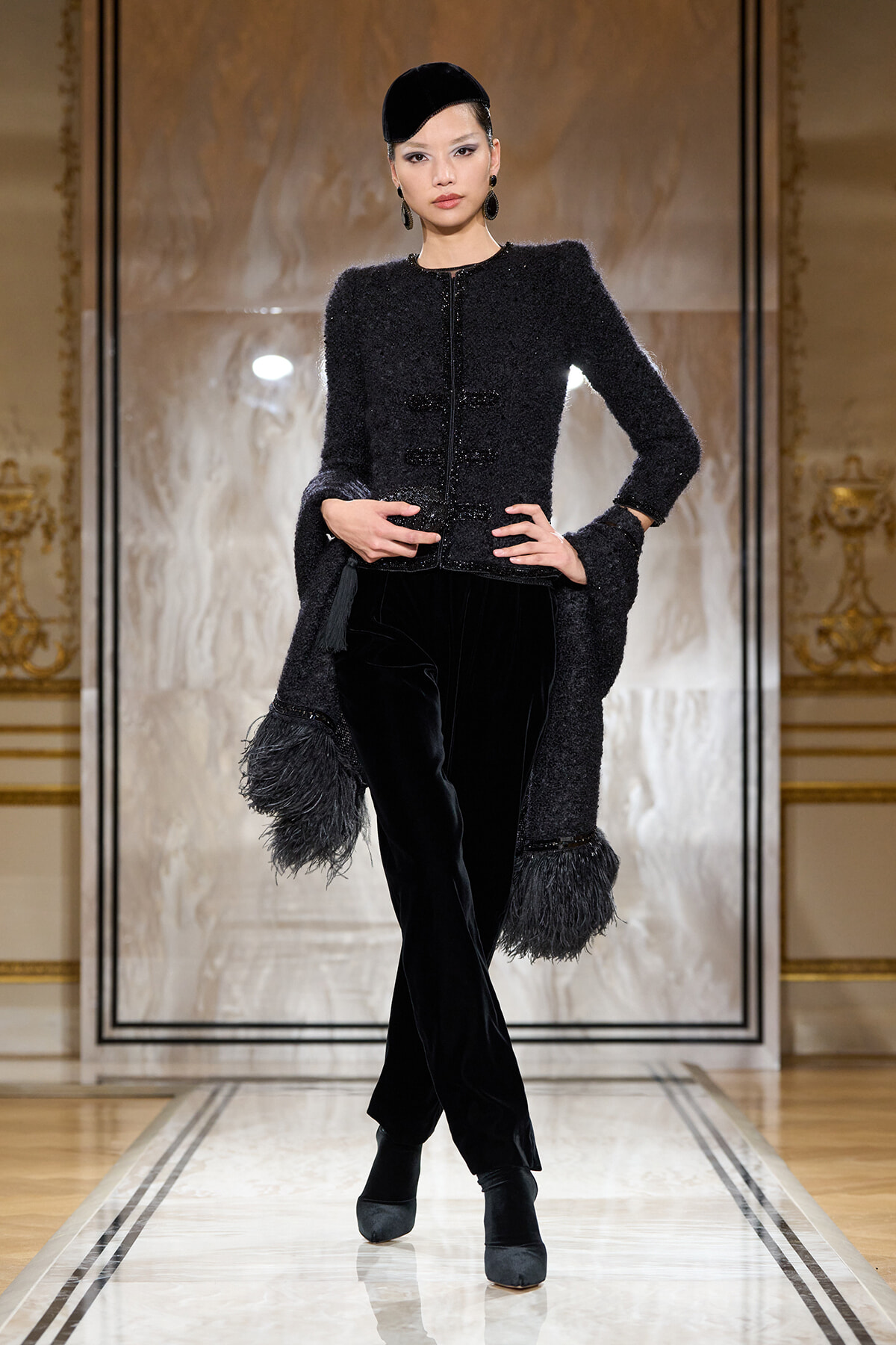 Model struts a runway in a black textured coat with fringe cuffs, matching velvet pants, and a small black cap.