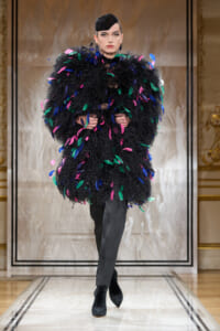 Model on a runway wearing a voluminous black feathered coat with multicolored leaf-shaped accents, paired with black pants and a sleek black hat.