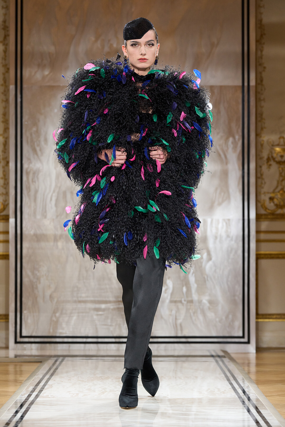 Model on a runway wearing a voluminous black feathered coat with multicolored leaf-shaped accents, paired with black pants and a sleek black hat.
