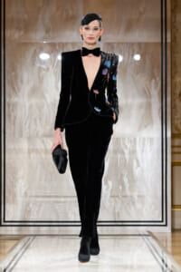 Model walks the runway in a black velvet tuxedo with colorful floral embroidery, deep V-neck, bow tie, and a small black clutch in hand.