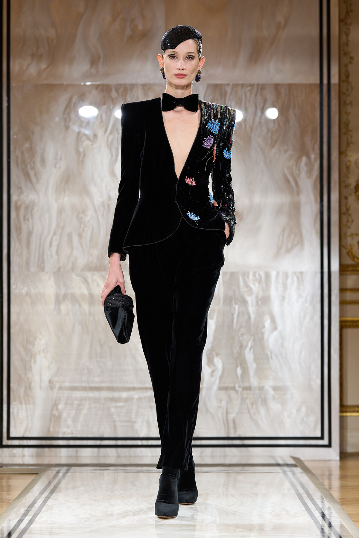 Model walks the runway in a black velvet tuxedo with colorful floral embroidery, deep V-neck, bow tie, and a small black clutch in hand.