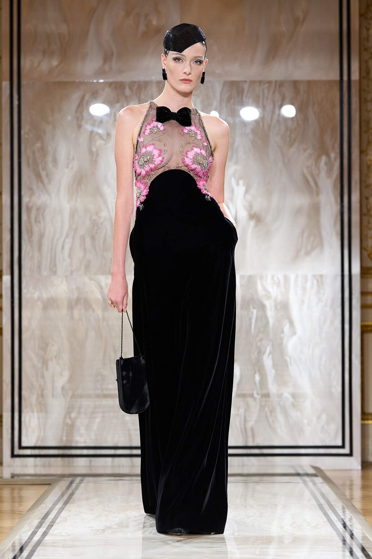 Runway model in a floor-length black velvet gown with a sheer pink-embroidered bodice and a small bow-tie detail.