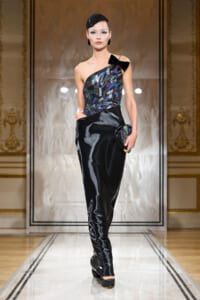 Model walking a fashion show in a black satin, one-shoulder gown with a multicolored sequin bodice and a black bow at the shoulder, carrying a small sequined clutch.