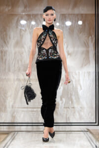 Model walks a runway in a black velvet pantsuit with an embellished halter top and bow, carrying a beaded black handbag.