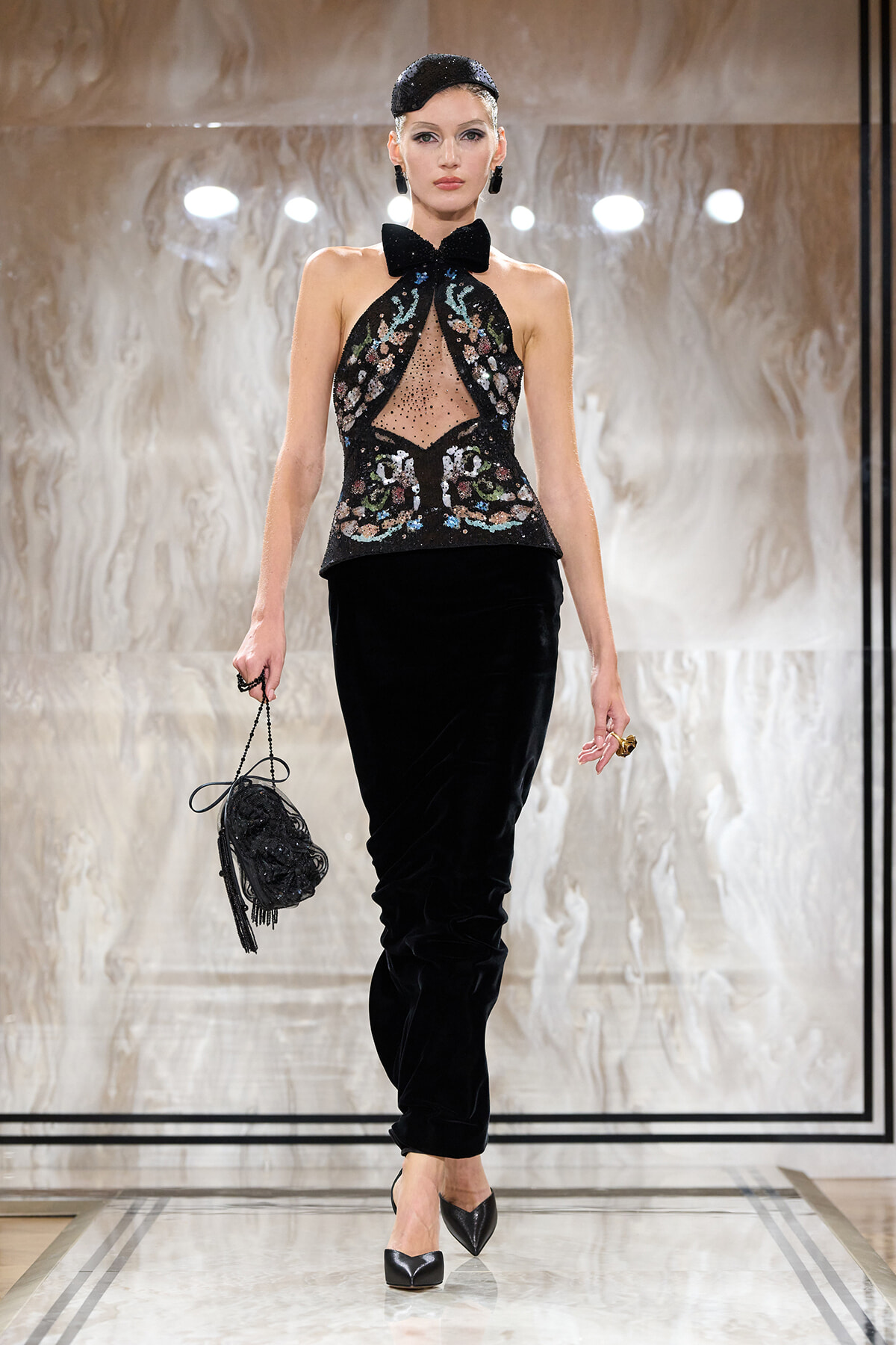 Model walks a runway in a black velvet pantsuit with an embellished halter top and bow, carrying a beaded black handbag.