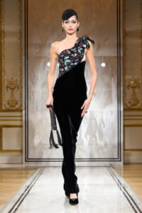 Model walking a runway in a black velvet one-shoulder gown with a sparkling floral-embellished bodice and a bow detail, accessorized with a black headpiece, statement earrings, and a beaded clutch with tassels.