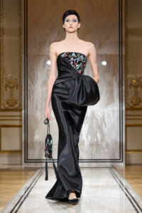 Model walks the runway in a strapless black satin gown with a colorful embroidered bodice and a large bow at the waist, carrying a beaded clutch.