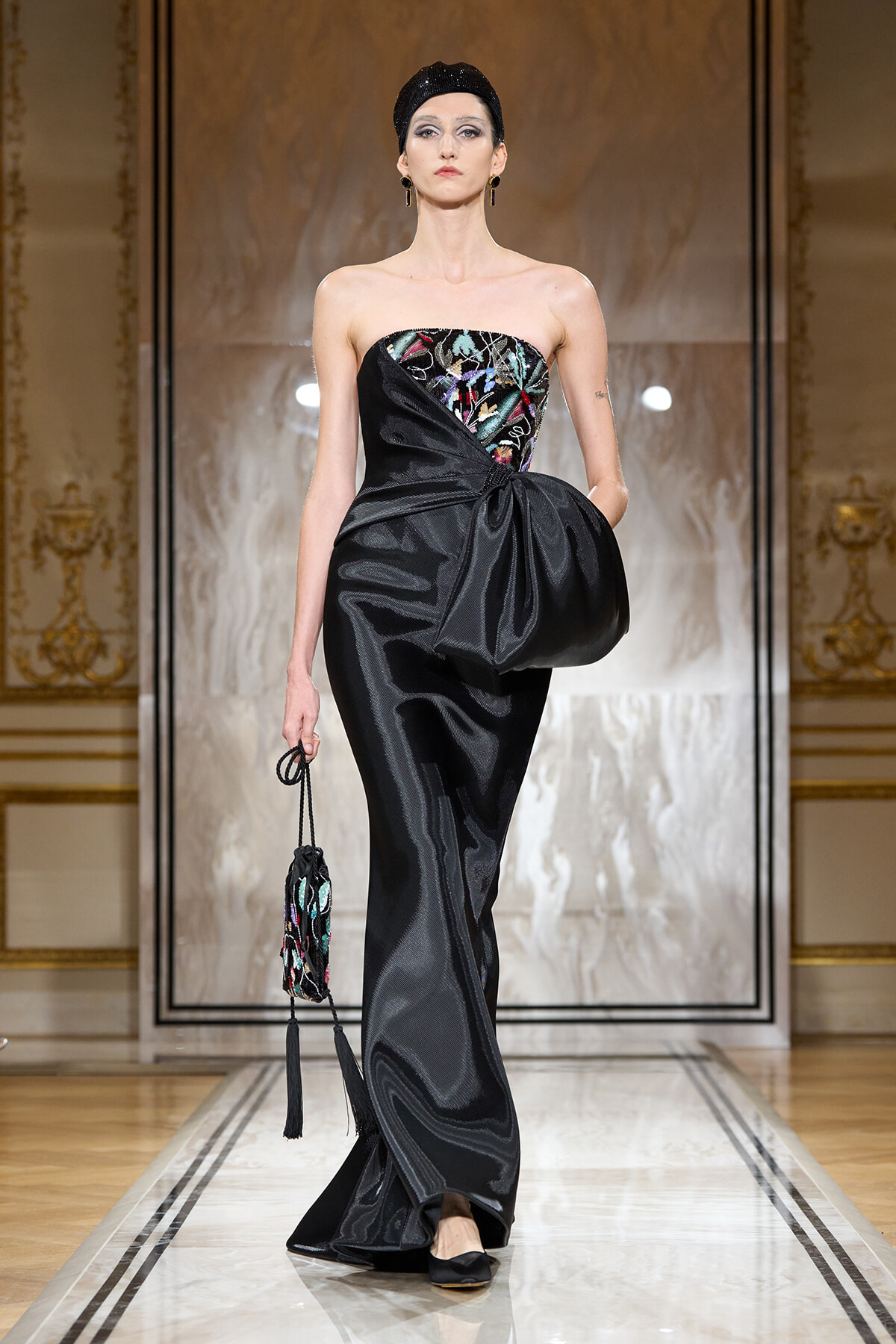Model walks the runway in a strapless black satin gown with a colorful embroidered bodice and a large bow at the waist, carrying a beaded clutch.