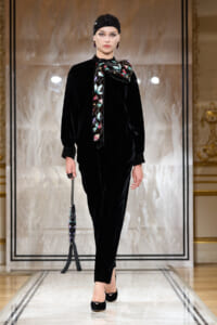 Model on a runway wearing a black velvet ensemble with a colorful embroidered bow scarf, matching headpiece, and a tasseled handbag.