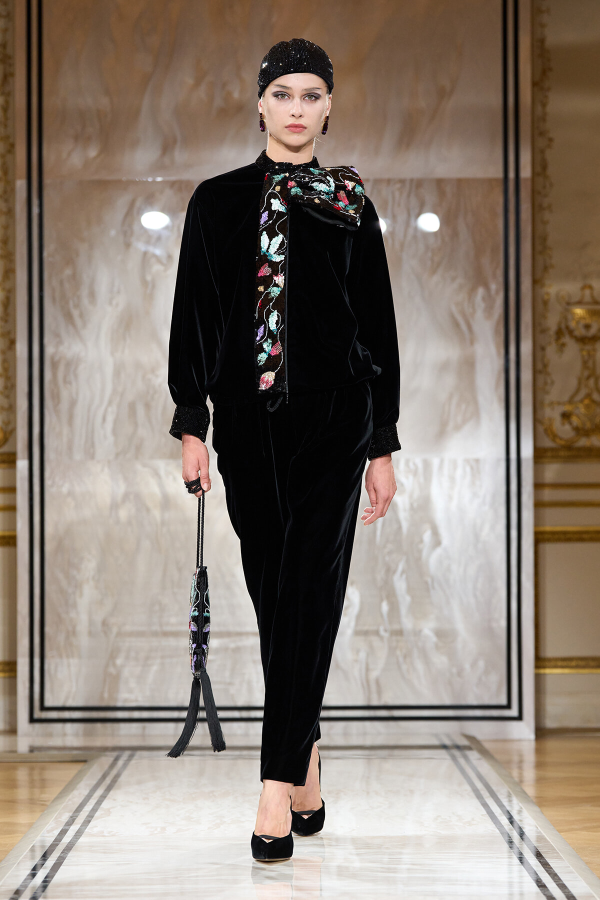 Model on a runway wearing a black velvet ensemble with a colorful embroidered bow scarf, matching headpiece, and a tasseled handbag.
