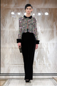 Model on a runway wearing a sequined multi-color cropped jacket with a black velvet collar over black wide-leg pants, paired with a black cap and clutch.