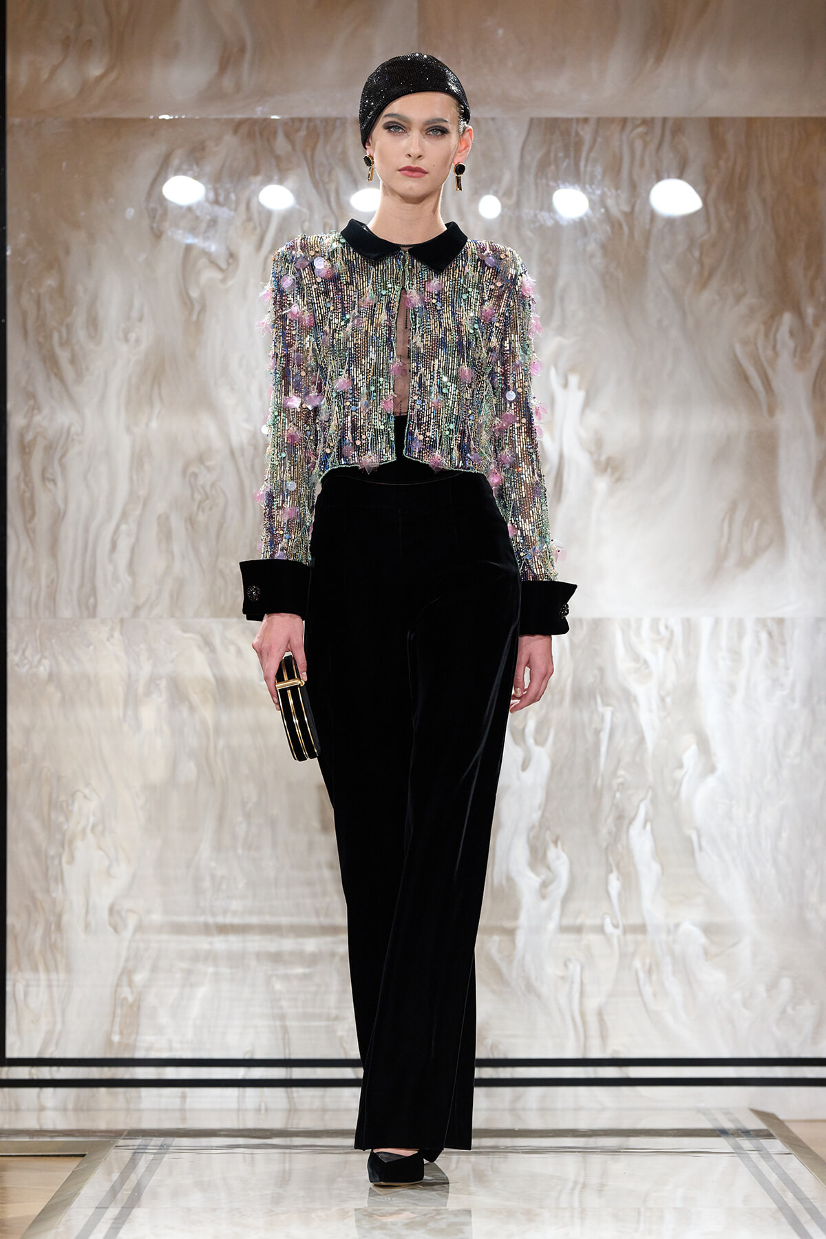 Model on a runway wearing a sequined multi-color cropped jacket with a black velvet collar over black wide-leg pants, paired with a black cap and clutch.