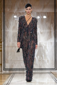 Model on a runway wearing a sheer, sequined gown covered in black pom-poms with a matching headpiece.