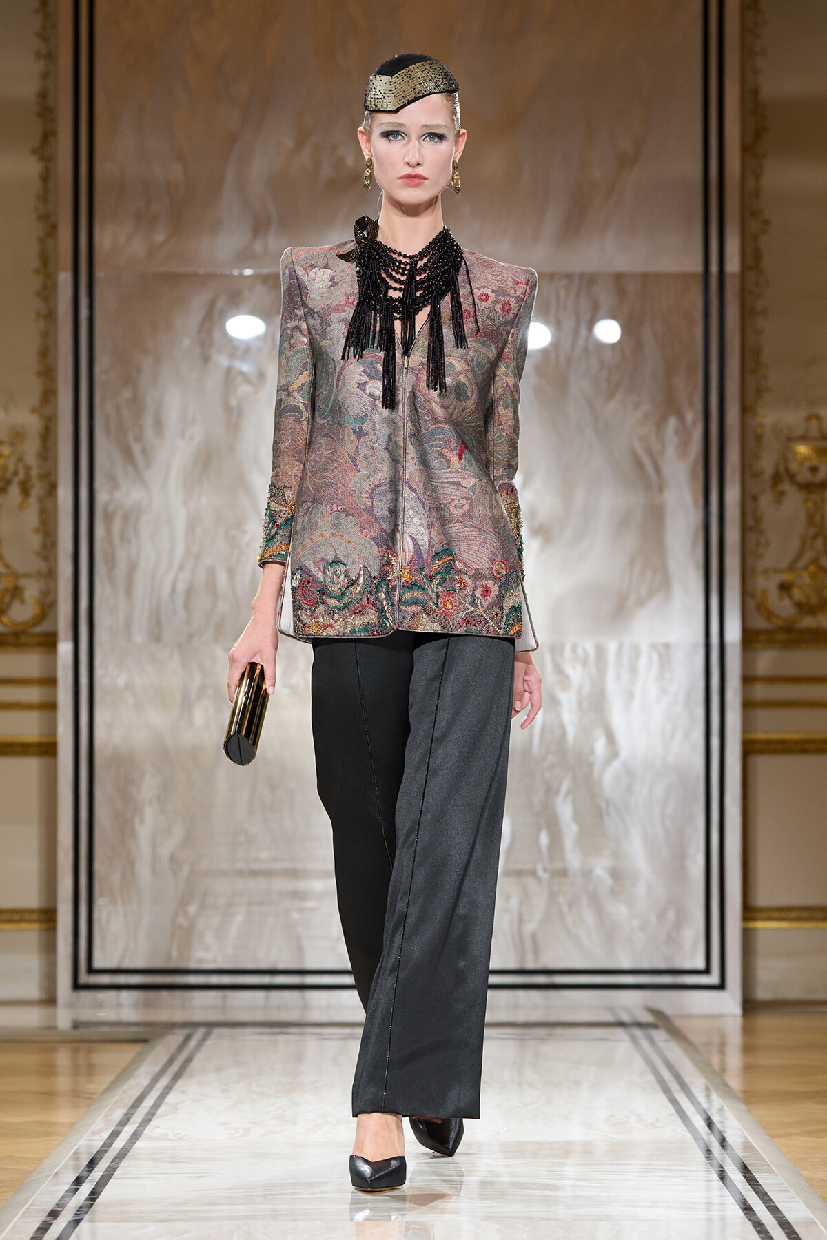 Model on runway wearing a metallic floral blazer, gray wide-leg pants, a bold black fringe necklace, a gold headpiece, and a gold clutch.