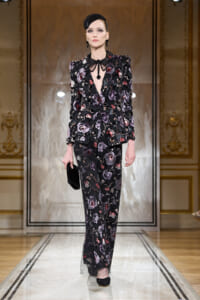 Model on a runway wearing a black floral suit with a deep V-neck, statement necklace, and black headpiece, carrying a black clutch.