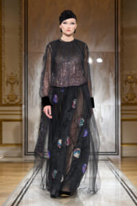 Model walks the runway in a black sequined top with a sheer tulle overlay and a long black skirt with colorful patch appliqués, wearing a black sequined cap.