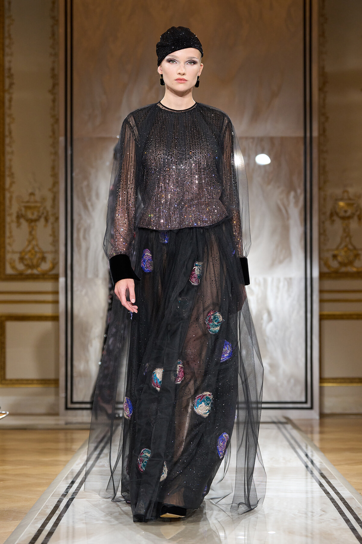 Model walks the runway in a black sequined top with a sheer tulle overlay and a long black skirt with colorful patch appliqués, wearing a black sequined cap.