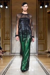 Model on a runway wearing a black beaded mesh cape over a black corset with green metallic wide-leg pants and a small black cap headpiece.