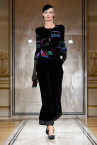 Model walking a luxurious runway in a black velvet suit with colorful geometric panels, paired with wide-leg velvet pants and gray cuffs, holding a small striped clutch.