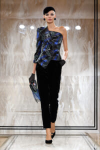 Model walking a runway in a one-shoulder blue-green patterned top and black velvet pants, paired with a black beret, gloves, and a fringe clutch.