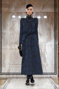 Model on a runway wearing a navy, textured long coat with a waist belt and high lace collar, paired with black gloves and a small black hat.