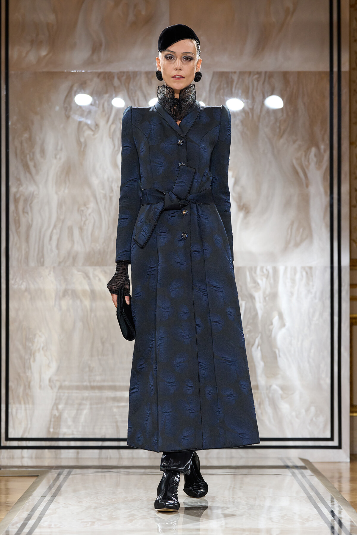 Model on a runway wearing a navy, textured long coat with a waist belt and high lace collar, paired with black gloves and a small black hat.