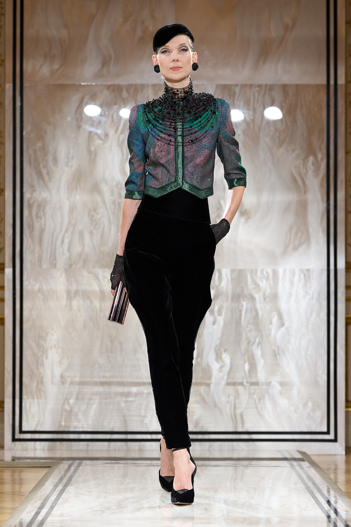 Runway model wearing a jeweled, green-embellished jacket with a high beaded collar, black velvet pants, black cap, gloves, and a striped clutch.