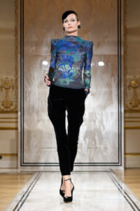 Model walking a runway in a metallic blue‑green patterned top with structured shoulders and black velvety pants, holding a dark clutch.