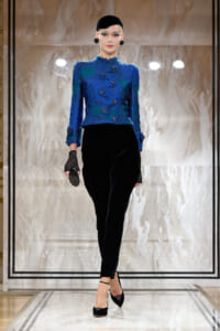 Model walking the runway in a blue embroidered jacket paired with black velvet pants and black accessories.